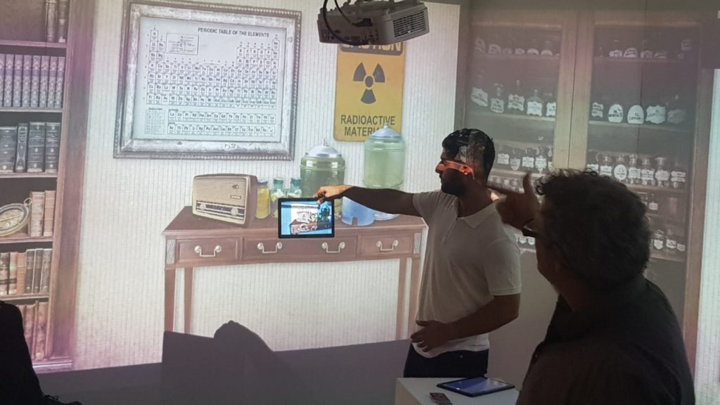 Foto de un hombre que sostiene una tableta frente a una escena proyectada de un laboratorio victoriano, con una tabla periódica, un cartel de materiales radiactivos y estantes con botellas de vidrio, mientras otro participante observa; muestra el uso de una tableta junto a la proyección en la pared