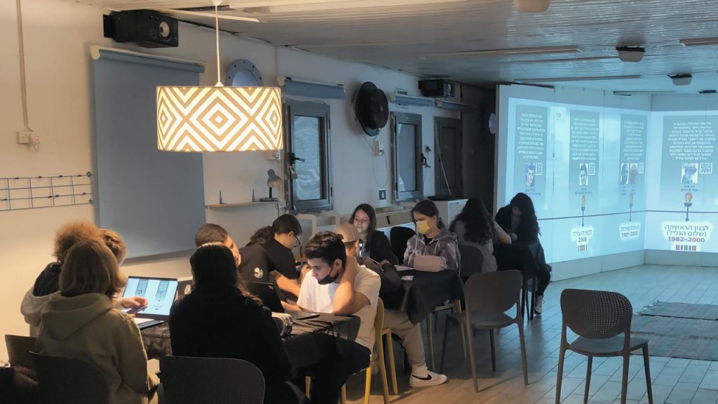 Foto de estudiantes que trabajan con portátiles en mesas mientras se proyecta, en las tres paredes de detrás, una cronología en hebreo de las campañas de Israel en el Líbano, con los periodos de 1982 a 2000 y de 1997 a 2003