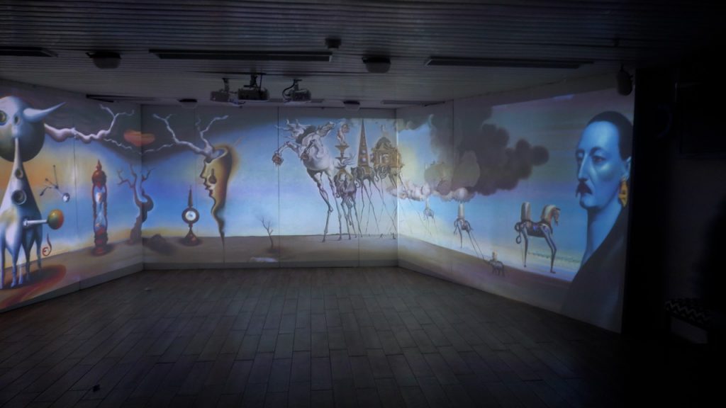Fotografía de una sala inmersiva de tres paredes con una panorámica surrealista al estilo de Dalí —relojes derretidos, criaturas de patas largas y delgadas y una cabeza masculina de perfil— proyectada sobre las paredes; se ven los proyectores del techo y el suelo de tablas de madera