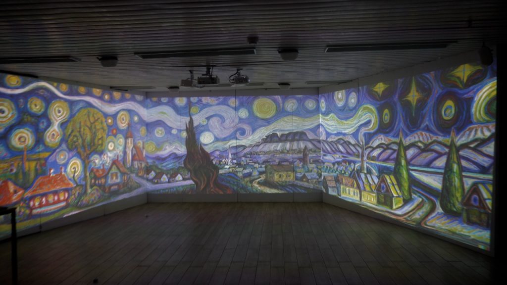 Fotografía de la misma sala inmersiva de tres paredes con una panorámica al estilo de «La noche estrellada» de Van Gogh —un cielo arremolinado, un pueblo de casas con tejados rojos y un ciprés alto y oscuro— proyectada sobre las paredes; se ven los proyectores del techo