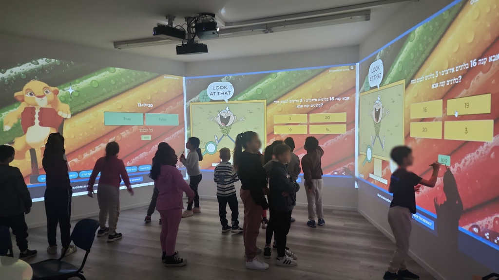 Foto de una clase de pie en la sala inmersiva durante una actividad de matemáticas: un problema en hebreo sobre globos con opciones de respuesta ocupa las paredes central y derecha, un cachorro animado llena la pared izquierda y un estudiante levanta la mano para tocar una respuesta