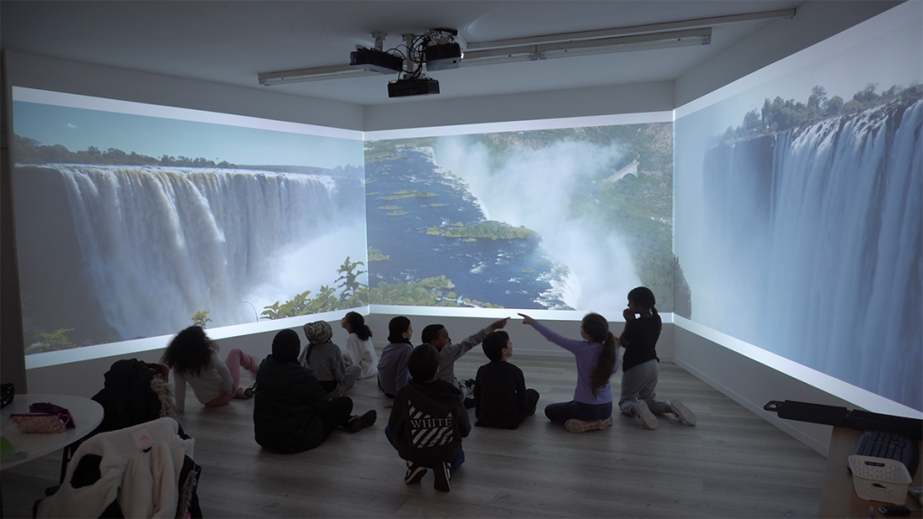 Foto de niños sentados en el suelo de la sala inmersiva frente a una panorámica de las cataratas Victoria que se extiende de forma continua por las tres paredes, con un niño señalando el agua que cae